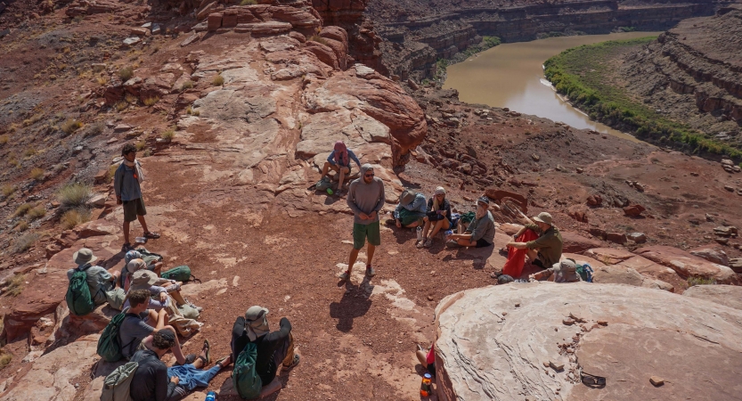 A group of students sit on the ground while listening to an instructor provide information. Below them, a river winds through canyon walls.